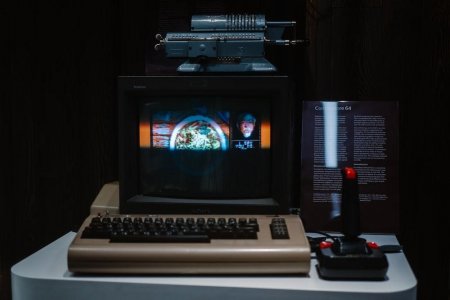 Auf einem Tisch steht ein Commodore 64. Auf dem Bildschirm ist ein farbiges Bild dargestellt. Neben der großen Tastatur steht ein Joystick.