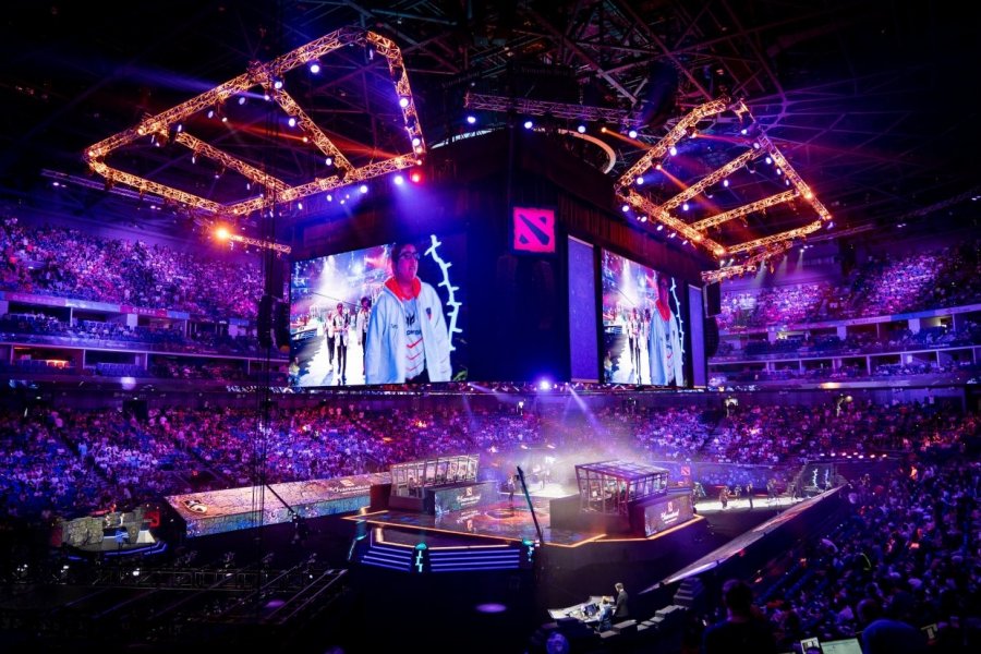Zu sehen ist eine mit Menschen gefüllte Arena. In der Mitte ist eine Bühne mit Computern. Auf großen Bildschirmen über der Bühne wird gezeigt, wie die Spieler in die Arena einlaufen.
